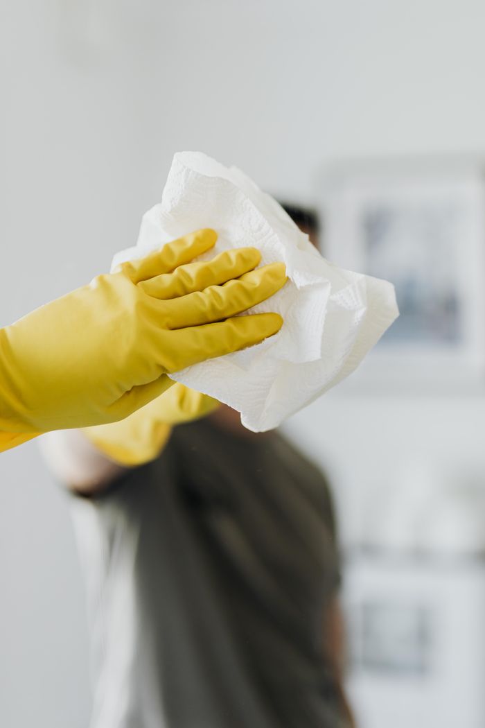 Homme anonyme portant des gants en latex jaunes et un t-shirt foncé, essuyant un miroir dans une salle de bain avec une serviette en papier